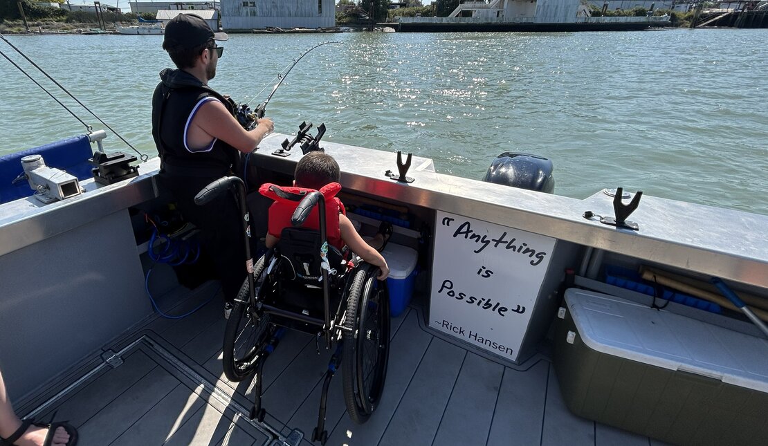 Person using a wheelchair on a boat deck holding a fishing rod beside another person, with water visible ahead and a sign reading, “Anything is possible.”