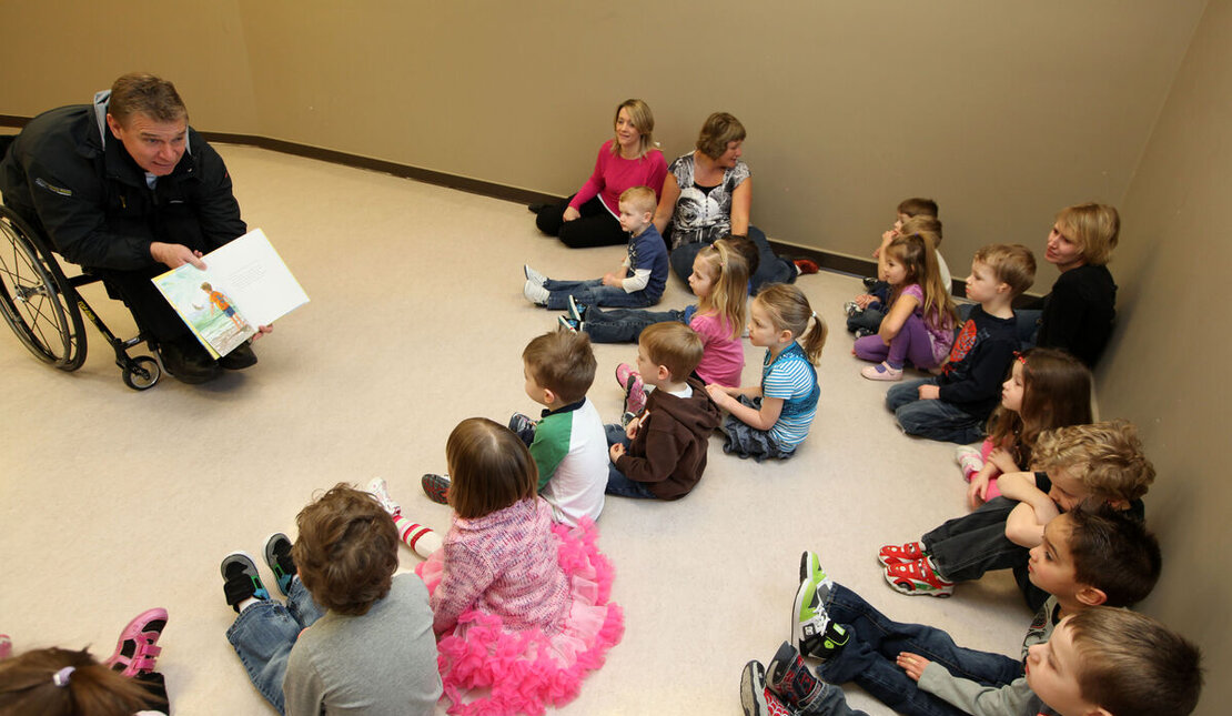 Man using a wheelchair (Rick Hansen) reading a book and showing its ilustrations to a group of children in a room