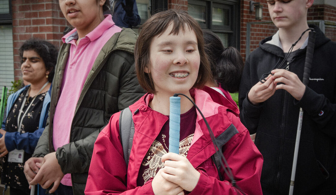 Group of young people standing outdoors, each holding a white cane. In the center, a young person with short dark hair wearing a red jacket holds a cane upright, with others nearby also holding canes in front of a brick building.