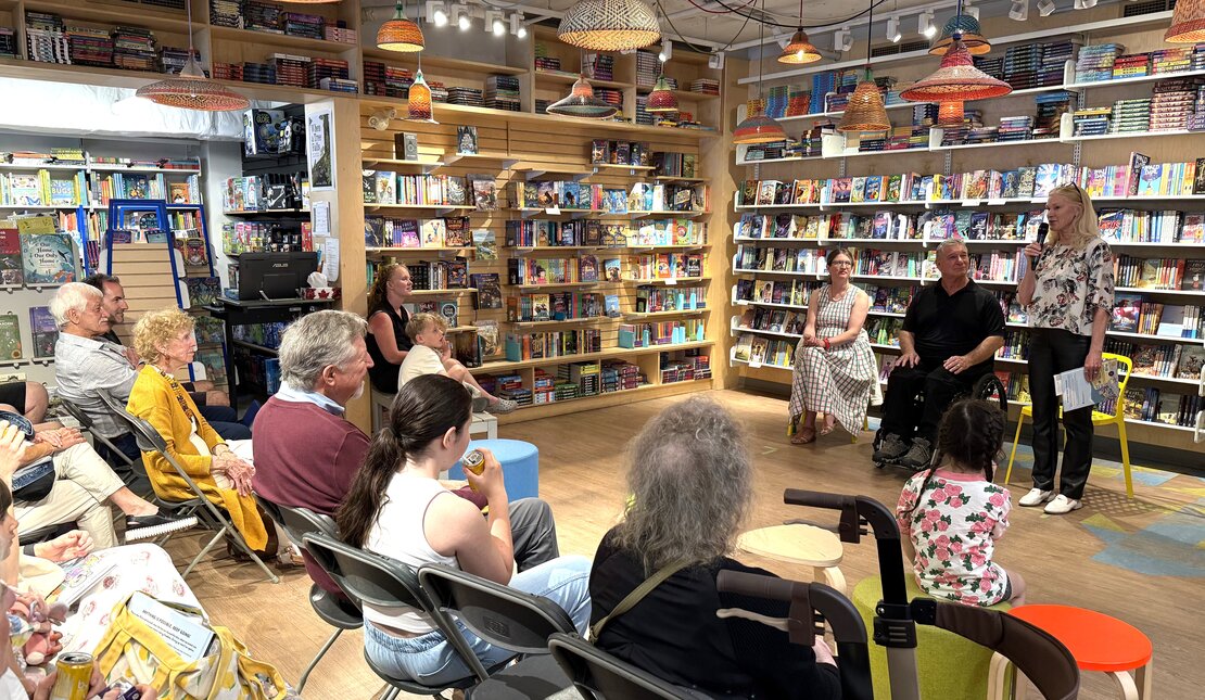 Bookstore event with an audience seated among bookshelves. At the front, three people sit and Lorna stands speaking into a microphone, with Rick and Emma the ilustrator seated among them.