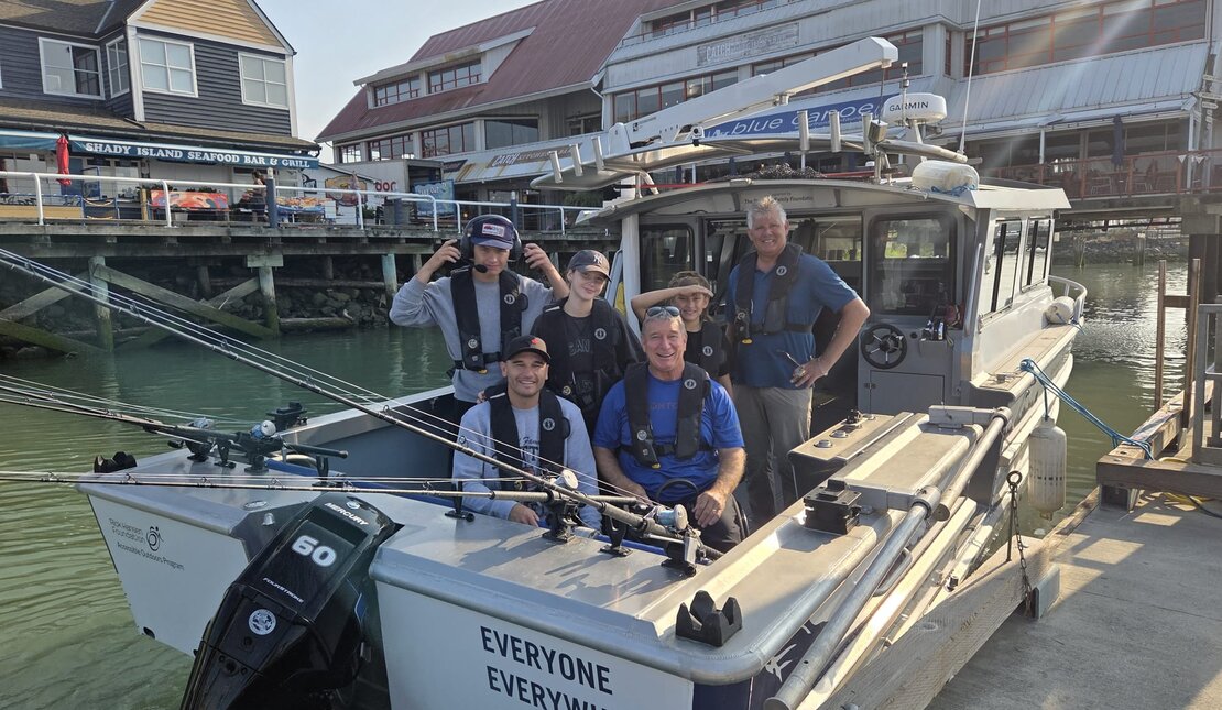 Group of people wearing life jackets aboard an accessible fishing boat docked at a marina. Fishing rods are mounted along the side, and waterfront buildings are visible in the background.