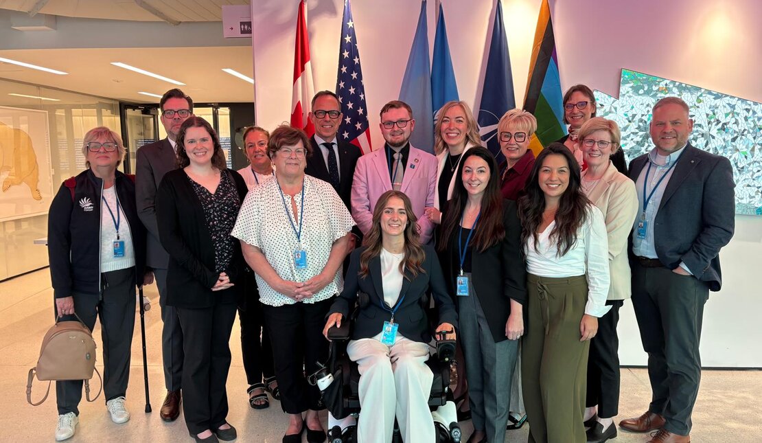 Group photo of delegates standing together indoors in front of several international flags. A young woman with long wavy brown hair is seated in a powered wheelchair at the center of the group, wearing a dark blazer and light-colored pants.