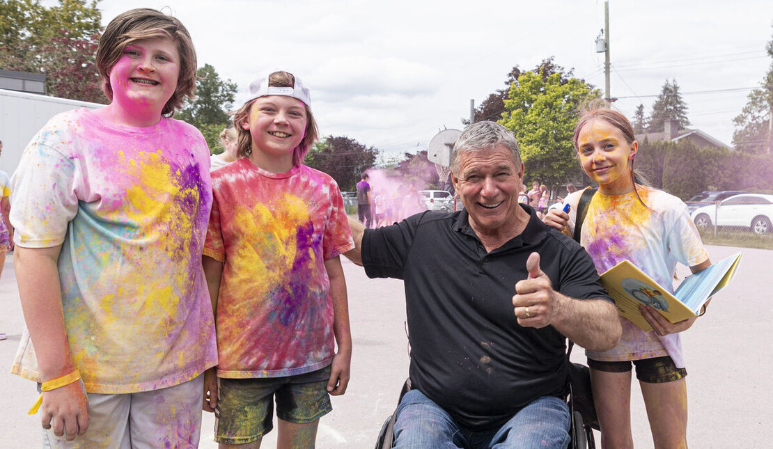 Three children covered in colorful powder stand outdoors beside Rick. One child holds an open book while the group poses on a school playground.