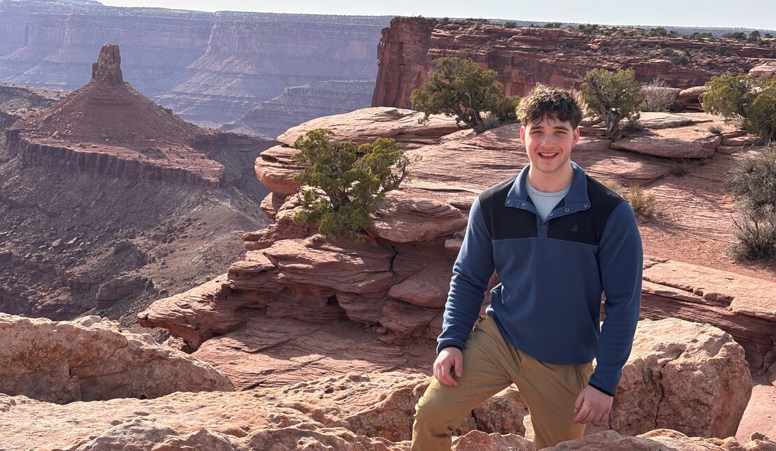 Young man with short, dark curly hair wearing a blue pullover and tan pants, standing among red rock formations overlooking a vast desert canyon with layered cliffs and scattered shrubs in the background.