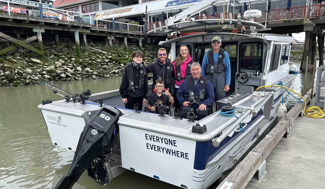 A group of five people aboard an accessible fishing boat docked at a marina. A man and a boy are using wheelchairs and are positioned near the centre of the group.