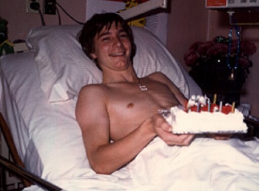 Rick Hansen smiles as he lays on hospital bed with cake during his 16th birthday