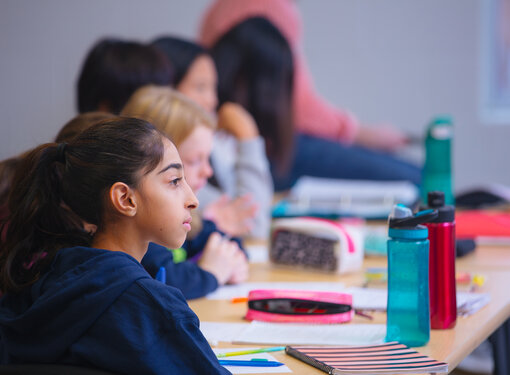 Side profile of a female elementary school student sitting at a desk and looking at the front of the classroom. The background is blurred, and includes a few of her classmates. Cover for Difference Maker (Grades K-8) toolkit.