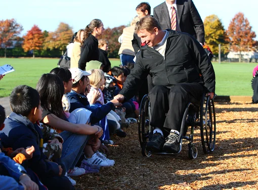 Rick Hansen wearing a black jacket, and in his wheelchair, bending over to shake hadns with one of many students that are sitting in a line on the ground outside. In the background is a school field and trees that have started changing colour in the autumn. Cover for Rick Hansen Story toolkit.