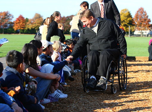 Rick Hansen wearing a black jacket, and in his wheelchair, bending over to shake hadns with one of many students that are sitting in a line on the ground outside. In the background is a school field and trees that have started changing colour in the autumn. Cover for Rick Hansen Story toolkit.