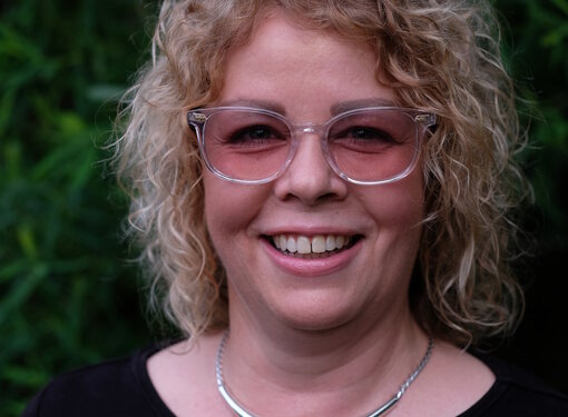 Woman with curly blonde hair, wearing pink sunglasses, smiling, wearing black blouse with necklace. 