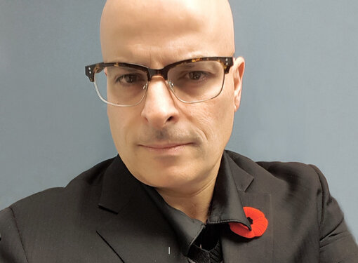 Middle aged bald man, brown eyes, wearing glasses, in a grey blazer. 