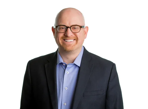 Bald man with blue eyes, wearing glasses, smiling in a dark grey blazer with blue shirt. 