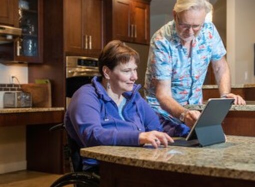 Woman using wheelchair and man standing use laptop in a kitchen on a low counter.