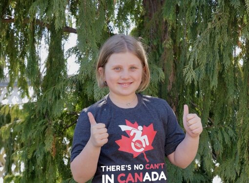 Young girl outdoors beside a tree, smiling with both thumbs up