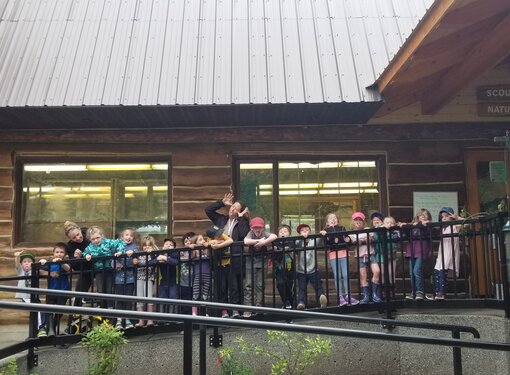 A group of children make funny faces with their teachers by the ramp railings outside Scout Island Nature Centre, a wooden structure with angled roof and and wide windows. 
