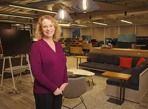 Liz Bailey Connor of First West Credit Union stands for a photo, she is wearing a purple camisole and has blond curly shoulder length hair. In the background are the furniture and lighting in the building.