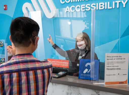 A man with a hearing device getting information from a female receptionist behind plexiglass. The woman is wearing a clear face mask and is pointing in the general direction beside her. There is a blue sign on the reception desk with a white icon that indicates the building is accessible for people with hearing loss.