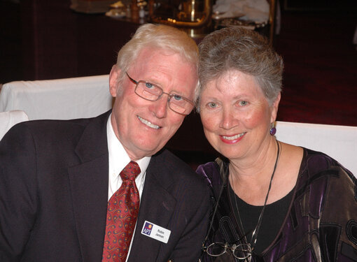 Robin and Cathryn sitting beside each other and smiling at the camera. They are older adults. Robin has white hair, glasses and is wearing a suit with a red tie. Cathryn has grey hair and is wearing purple earrings and a purple and black dress.