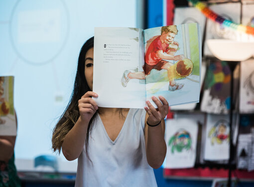 Person with long dark hair wearing a white t-shirt holding open a children's book with a colourful illustration.