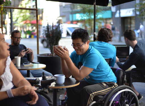 Man with black hair and glasses using a wheelchair wearing a bright blue t-shirt sitting in a cafe. He is holding a coffee cup.