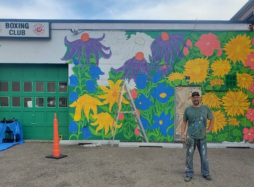Man with clothes covered in paint standing in front of a large, colourful flower mural.