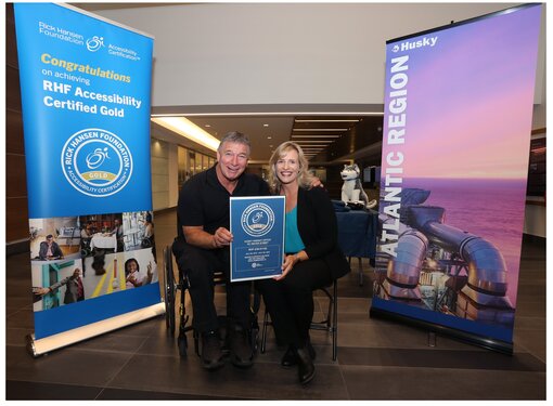 Rick Hansen (man with light brown hair) using his wheelchair holding a blue certification sign with a blonde woman who is sitting beside him. They are beside two large banners.