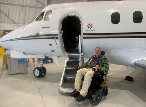 Man with light hair who is using a motorized wheelchair next to a plane. He is wearing light pants and a green and grey jacket.