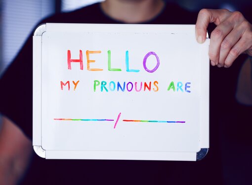 Person holding up a whiteboard wearing a black t-shirt. In rainbow marker, the sign reads hello, my pronouns are blank space slash blank space.