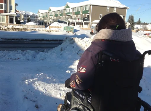 Back of person using a wheelchair wearing a purple winter jacket. It is winter with large piles of snow on the ground and apartment buildings in the distance. 