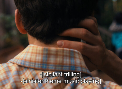 Back of the head of someone with short dark hair wearing a plaid shirt whose hand is on the back of their neck. Closed captioning located at the bottom of the image reads "sibilant trilling" and "sinister theme music playing".