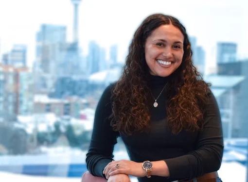 Haley Rae Dinnall-Atkinson, who is a woman with long brown curly hair smiling and leaning on a brown chair. She is wearing a black turtle neck sweater and blue jeans. Behind her is an open window to a city landscape.