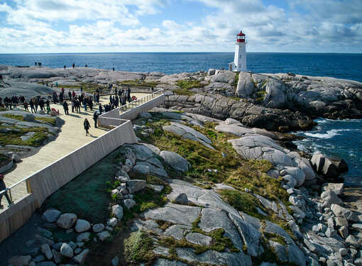 Aerial view of Peggy's Cove lighthouse. There is a large, wooden accessible viewing deck with a crowd of people towards the front. The viewing deck is built on top of large rocks. The lighthouse is red and white and overlooks the ocean.