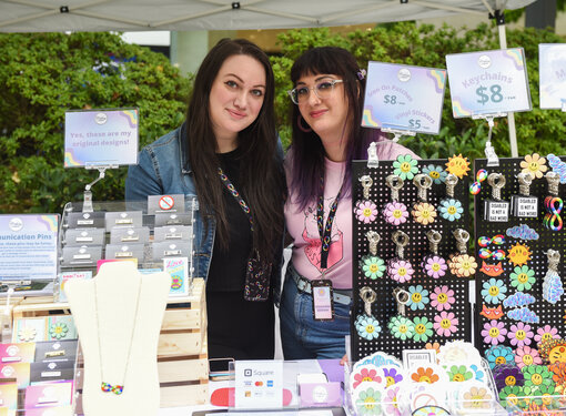 We Belong! Festival founder Margaux Wosk on the right with sister Becky Wosk. Margaux has long dark hair with purple in it, is wearing a pink t-shirt and has glasses. Becky has long dark hair and is wearing a black outfit with a denim jacket. They are in front of a stand of items that are for sale.