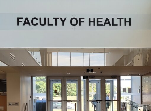 Inside the University of Waterloo. The Faculty of Health sign is above a set of doors with stairs and an accessible ramp.