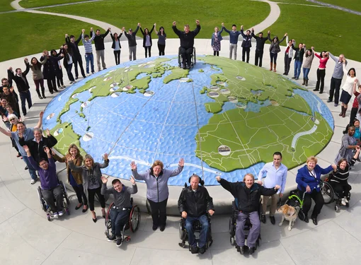 A group of people of different abilities around the perimeter of a large globe. They are celebrating. 