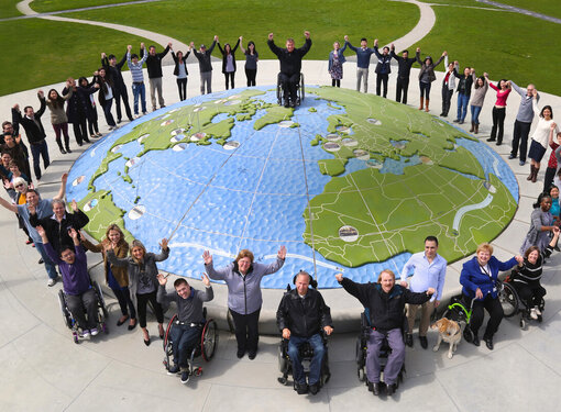 A group of people of different abilities around the perimeter of a large globe. They are celebrating. 