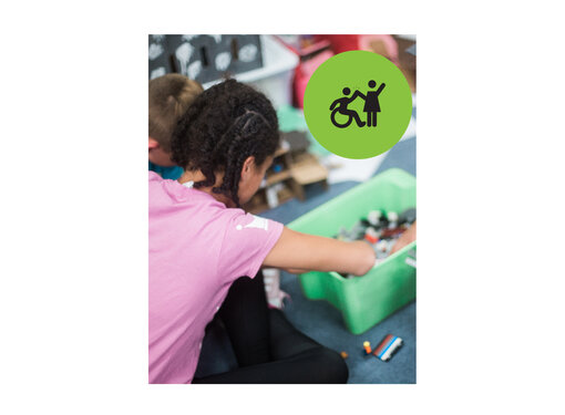 Girl wearing mauve shirt crouching and digging through a crate of lego. Small green circle icon with graphic of person in a wheelchair high-fiving a person that is standing.