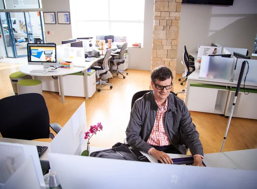 Person with short brown hair wearing a grey jacket working at a computer in an office. There is a white cane by the desk.
