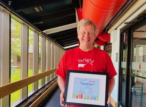 Archie who is a man with short blonde hair wearing a red t-shirt. He is holding a framed Difference Maker of the Year award certificate. 