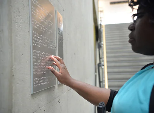 Woman wearing a teal shirt reading a metal braille sign that is mounted on a wall.