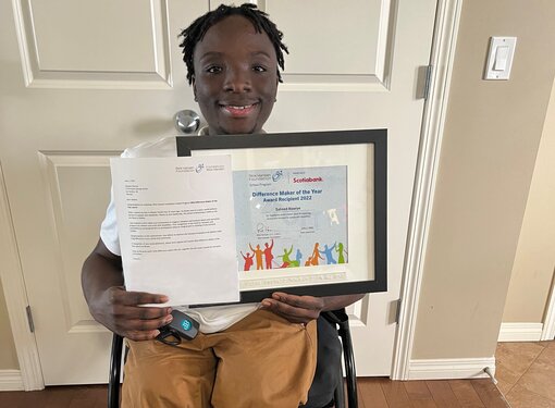 Saheed who is using his wheelchair holding his Difference Maker of the Year Award certificate. He is smiling and wearing tan-coloured pants.