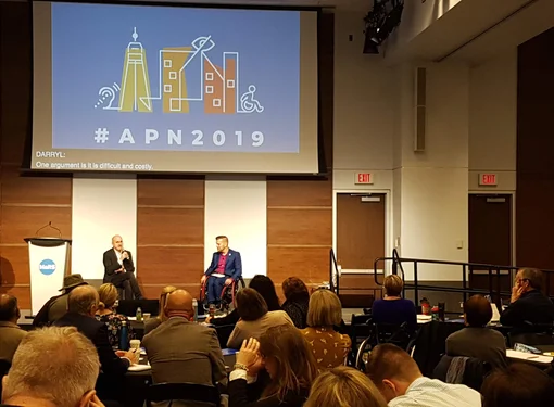 APN 2019. A conference room filled with people looking at two speakers at the front of the room. One of the speakers is using a wheelchair. There is an ASL interpreter and a projector that says #APN2019