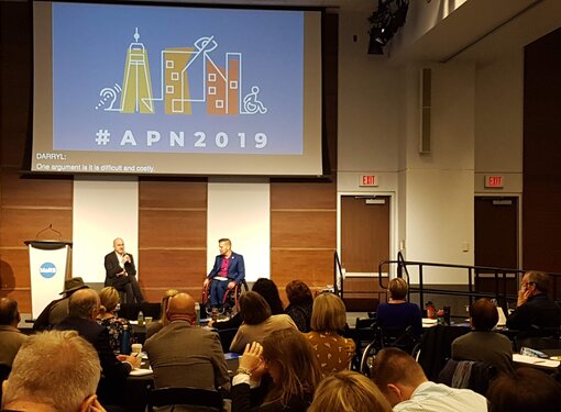 APN 2019. A conference room filled with people looking at two speakers at the front of the room. One of the speakers is using a wheelchair. There is an ASL interpreter and a projector that says #APN2019