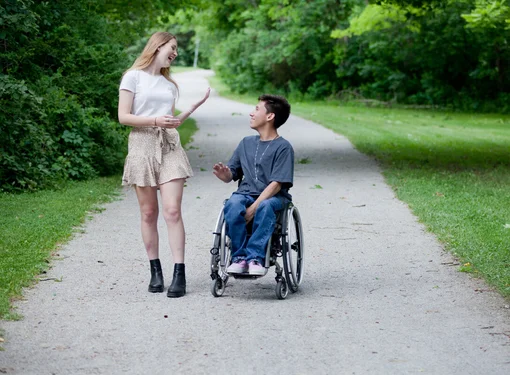 Two people talking and laughing while on a gravel path. One is using a wheelchair, has short brown hair and is wearing a dark t-shirt. The other person has long hair, is wearing a skirt and is walking.