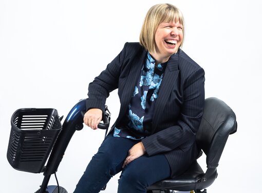 Darby Lee Young who has short blonde hair cut in a bob. Darby is wearing a striped blazer, a floral shirt and shiny blue shoes. She is laughing and is using a motorized mobility device.