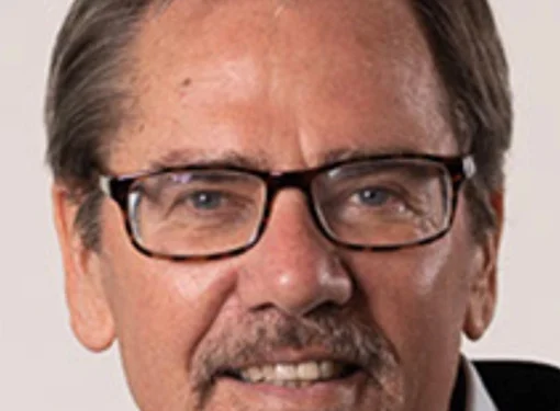 Person with short brown hair and a beard with glasses wearing a button up blue shirt and a black jacket.