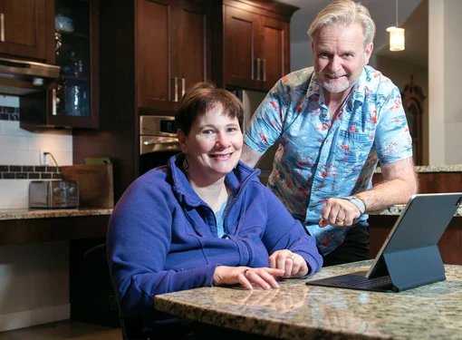 Couple at their kitchen counter which is at an accessible height. There is an iPad on the counter. The man has grey hair and is standing next to his wife who has short brown hair and is using a wheelchair.