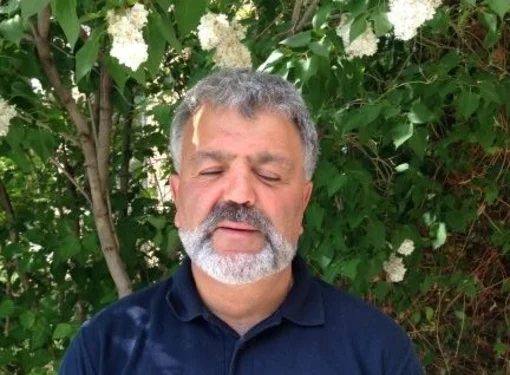 Lui Greco, who has salt and pepper hair and heard, is wearing a dark blue collared shirt and is blind. He is in front of trees.