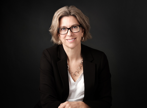 Headshot of Stephanie Cadieux who has short brown hair and glasses. She is wearing a black blazer and white shirt and is in front of a black background.