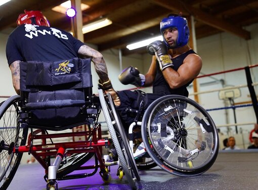 Two people who are using wheelchairs boxing in a ring. One person is wearing a red helmet and the other is wearing a blue helmet. 